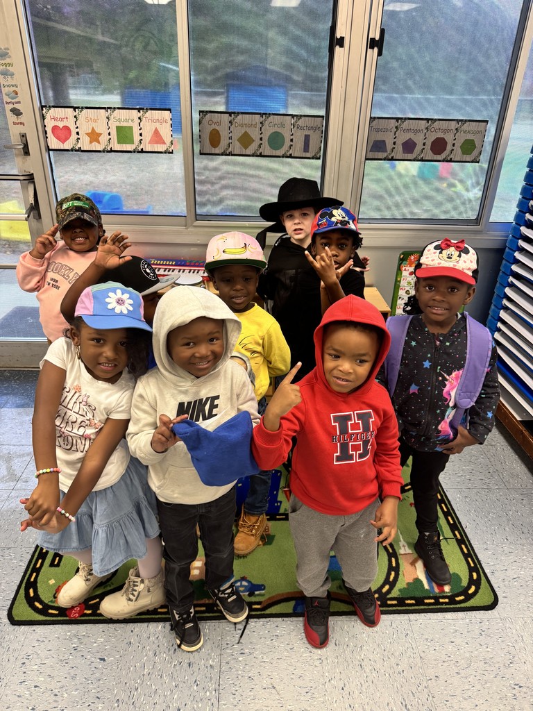 A group of students stand together on a colorful classroom rug, all wearing fun hats and hoodies. Some make peace signs while smiling for the camera in front of a bright classroom window.