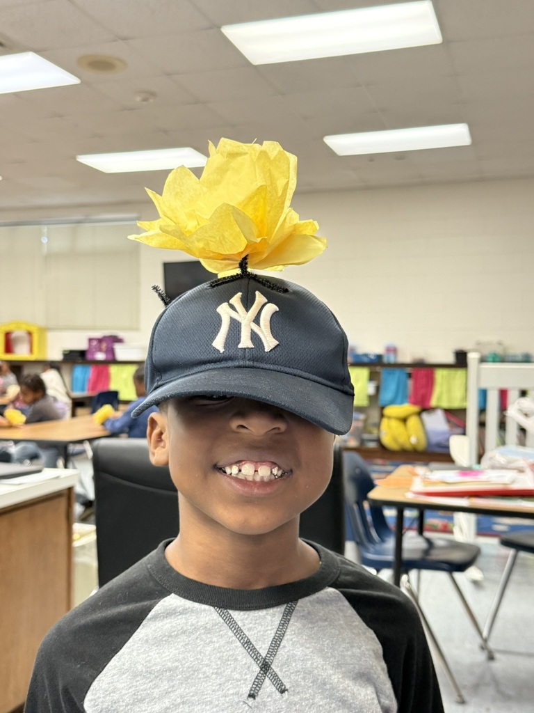 A smiling student wearing a dark gray shirt and a navy Yankees baseball cap has a bright yellow tissue paper flower attached to the top of his hat.