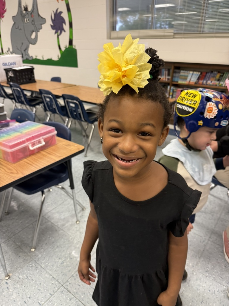 A smiling student wearing a black dress with a large yellow paper flower pinned in her hair stands in the classroom, with tables and chairs in the background.