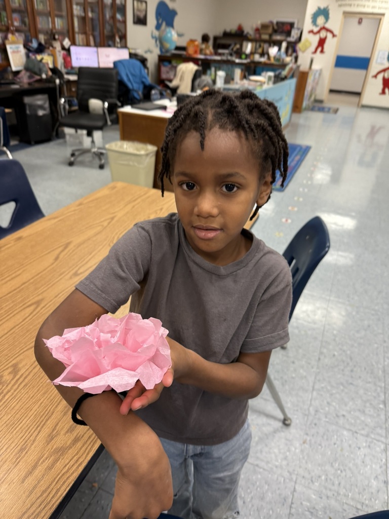 A student in a gray shirt shows off a pink tissue paper flower tied around his wrist while sitting in the library classroom.