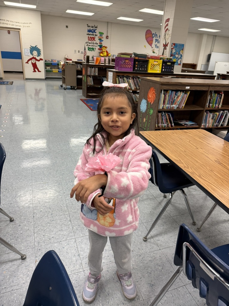 A smiling student in a pink fleece jacket stands in the library, showing her pink tissue paper flower bracelet.