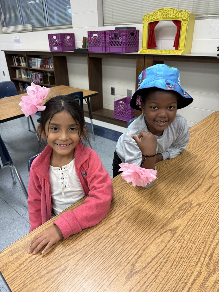 Two students sit side-by-side at a table, each with pink tissue paper flowers. One wears a pink fleece jacket and the other a blue bucket hat decorated with cartoon characters.