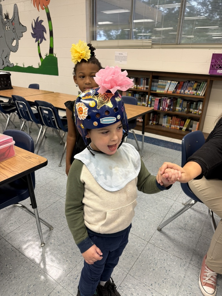 Two students are pictured—one wearing a yellow flower in her hair, and another smiling with a pink paper flower attached to a decorated blue helmet covered in stickers that say “New Driver” and “Caution.”