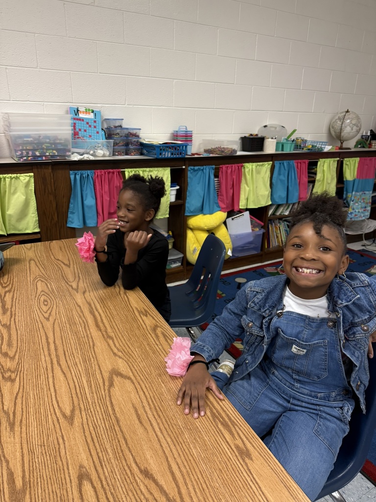 wo students sit at a classroom table smiling, each wearing pink tissue paper flowers on their wrists. One wears denim overalls and the other a black dress.