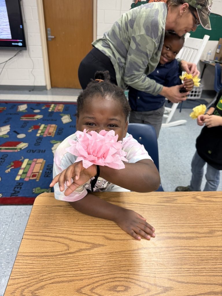 A student sitting at a table laughs as she holds a large pink paper flower to her face. In the background, another student is being helped by a teacher attaching a yellow flower.