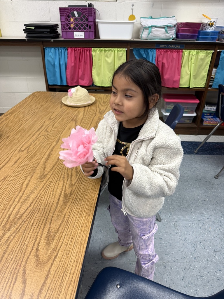 A young student in a light-colored jacket holds up a pink paper flower she made, smiling proudly at her work.