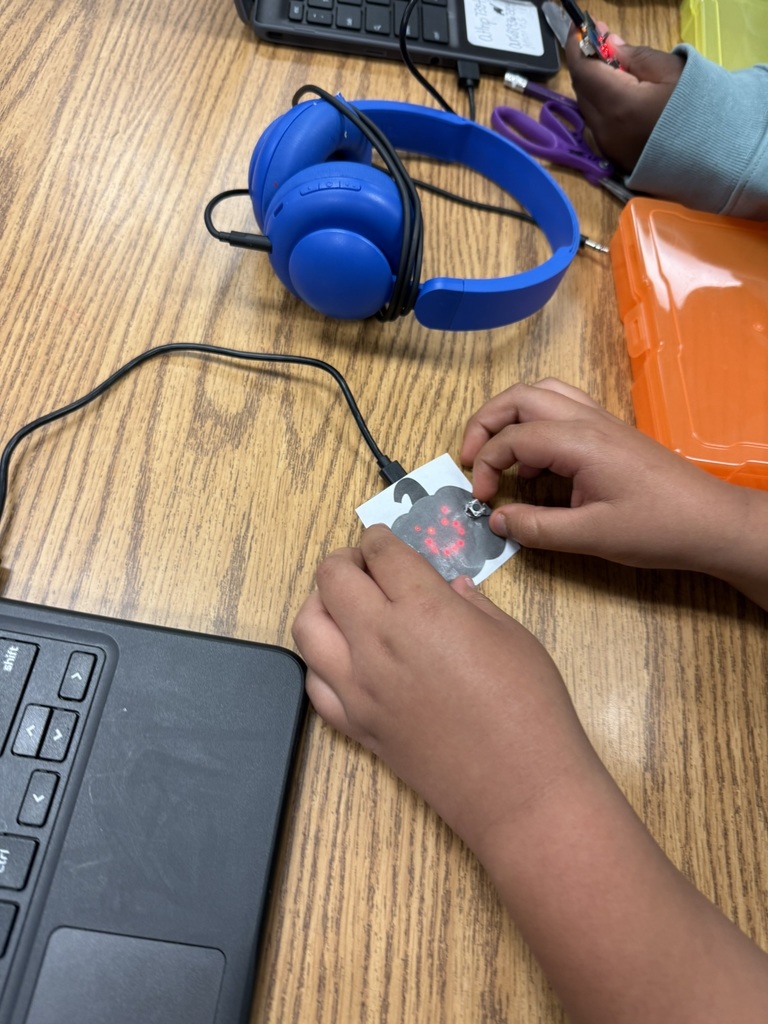 Hands of a student working with a micro:bit connected by a cable to a small paper pumpkin with red LED lights. Blue headphones and classroom supplies are visible on the table.
