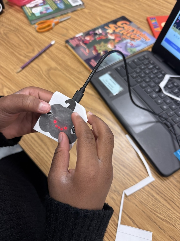 Close-up of a student’s hands holding a small paper pumpkin shape with a micro:bit attached, showing glowing red LED lights in a face pattern.