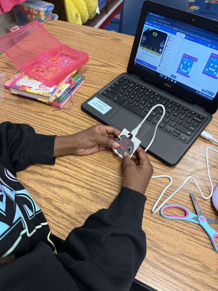 A student’s hands hold a micro:bit showing a red LED face design on a paper pumpkin while coding on a nearby laptop displaying block-based programming.