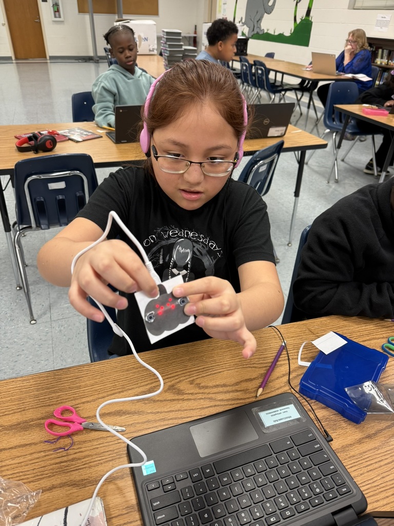 A student wearing pink headphones focuses on coding at her laptop while holding a small paper jack-o’-lantern connected by wires to a micro:bit device with red LED lights glowing.