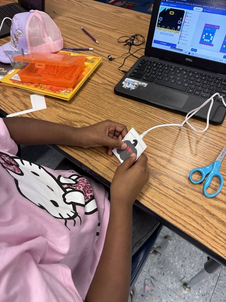 A student wearing a pink Hello Kitty shirt codes at her laptop while holding a small paper pumpkin powered by a micro:bit with glowing lights.