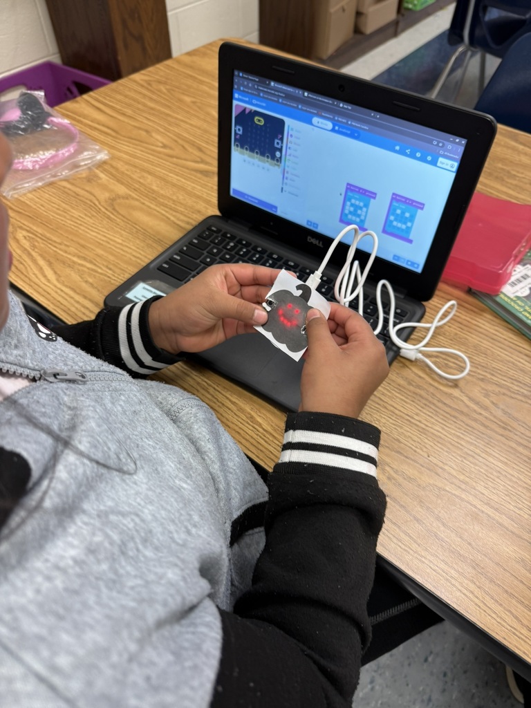 A student holds a micro:bit-powered paper pumpkin showing a red LED display while looking at coding blocks on her laptop screen.