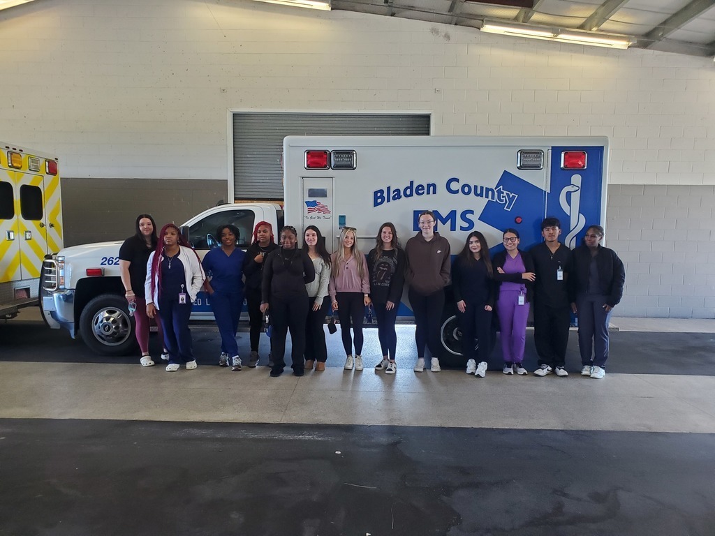 A group of thirteen people standing in front of a Bladen County EMS ambulance inside a garage. They are smiling and posing together, with another yellow-striped ambulance partially visible on the left.