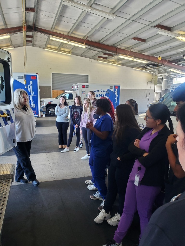 A group of students stands inside an EMS station, listening to an instructor beside an open ambulance. The instructor is explaining something about the vehicle, while multiple ambulances are visible in the background. The students appear attentive, some wearing scrubs, indicating a medical or emergency training session.