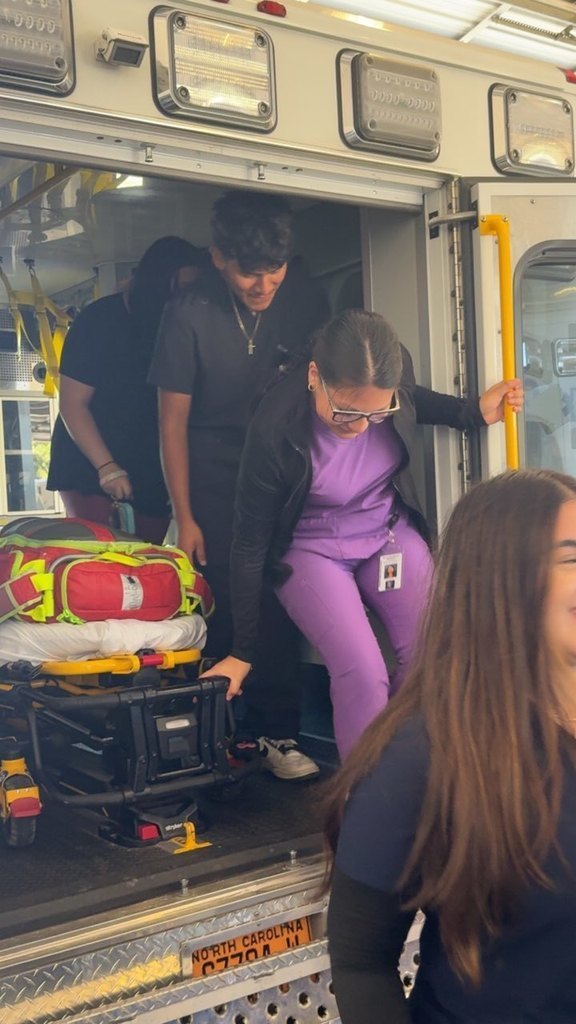 A group of students step out of the back of an ambulance. One person in purple scrubs is holding the yellow handle and stepping down carefully, while another in black scrubs follows behind. A stretcher with a red and green medical bag is visible inside the ambulance.