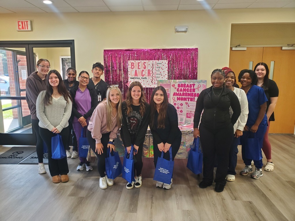 A group of students pose together indoors in front of a pink, sparkly Breast Cancer Awareness Month display board. They are smiling and holding blue tote bags. The background includes doors, windows, and a sign with pink decorations promoting breast cancer awareness.