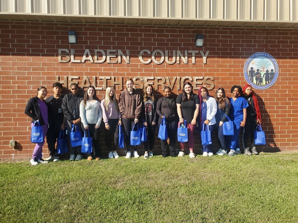 A group of students standing in a line outside the Bladen County Health Services building, holding blue tote bags. The building has a brick wall with the organization’s name and logo displayed.