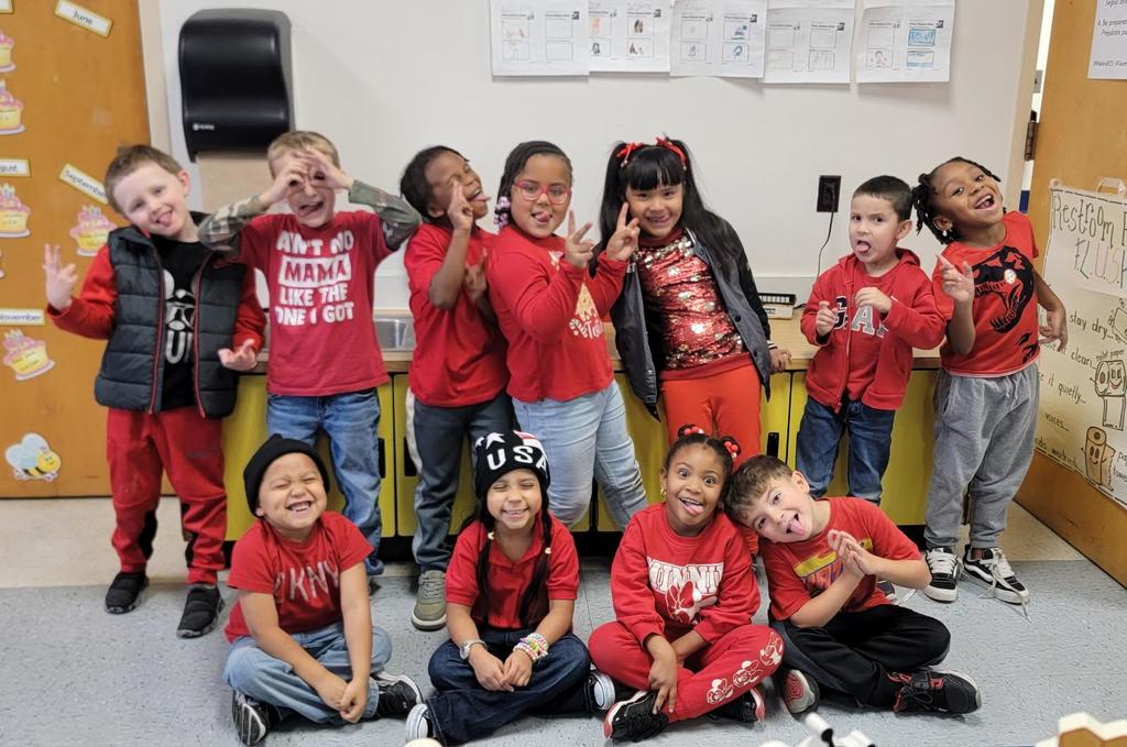 A group of young elementary students and two teachers pose together in a colorful classroom. Everyone is smiling and wearing red clothing to celebrate Red Ribbon Week.