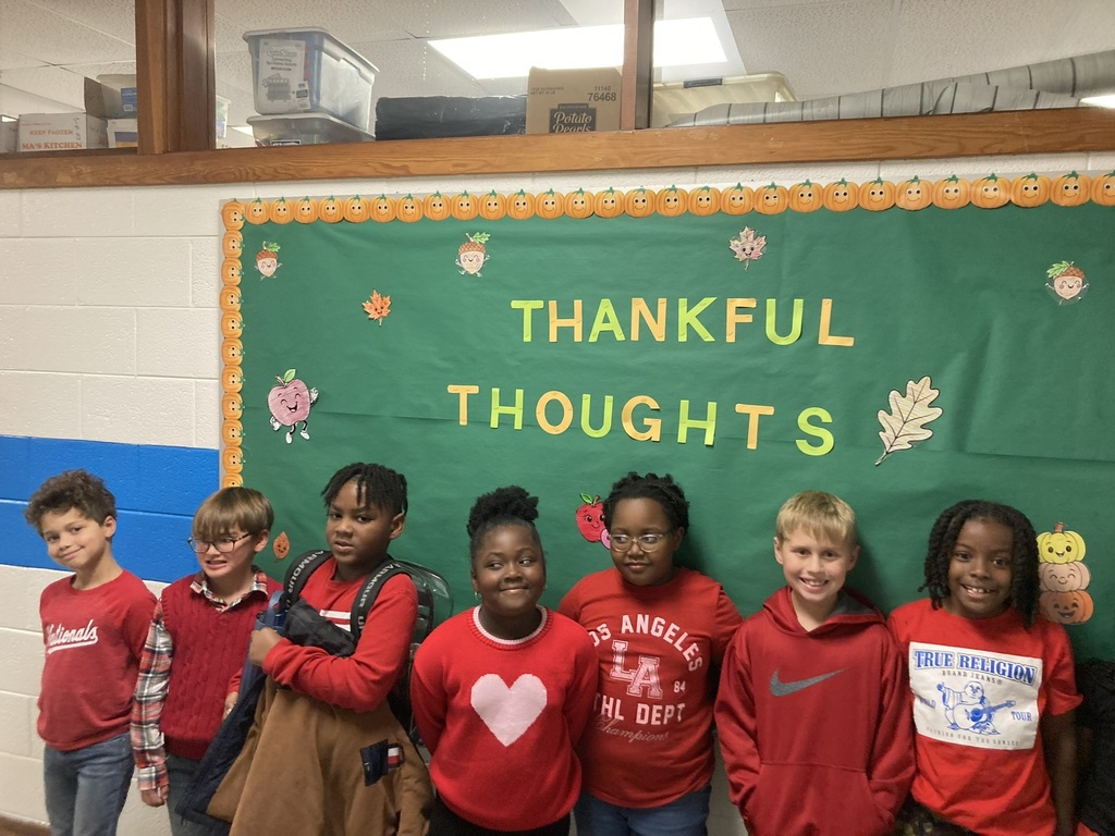 A group of young elementary students and two teachers pose together in a colorful classroom. Everyone is smiling and wearing red clothing to celebrate Red Ribbon Week.