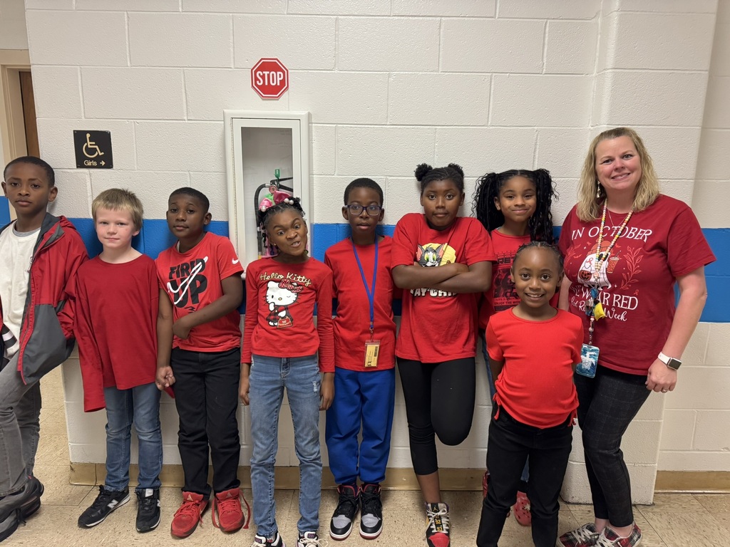 A group of young elementary students and two teachers pose together in a colorful classroom. Everyone is smiling and wearing red clothing to celebrate Red Ribbon Week.
