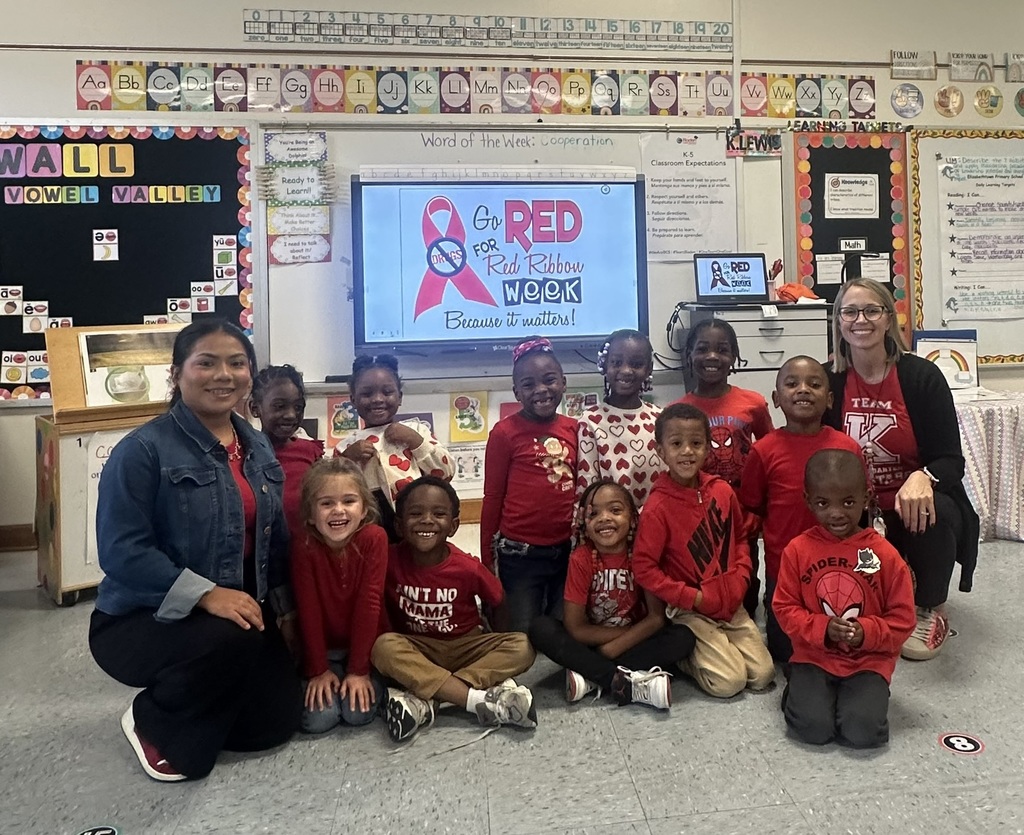 A group of young elementary students and two teachers pose together in a colorful classroom. Everyone is smiling and wearing red clothing to celebrate Red Ribbon Week.