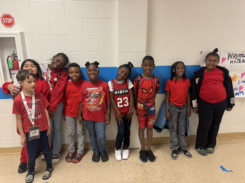 A group of young elementary students and two teachers pose together in a colorful classroom. Everyone is smiling and wearing red clothing to celebrate Red Ribbon Week.