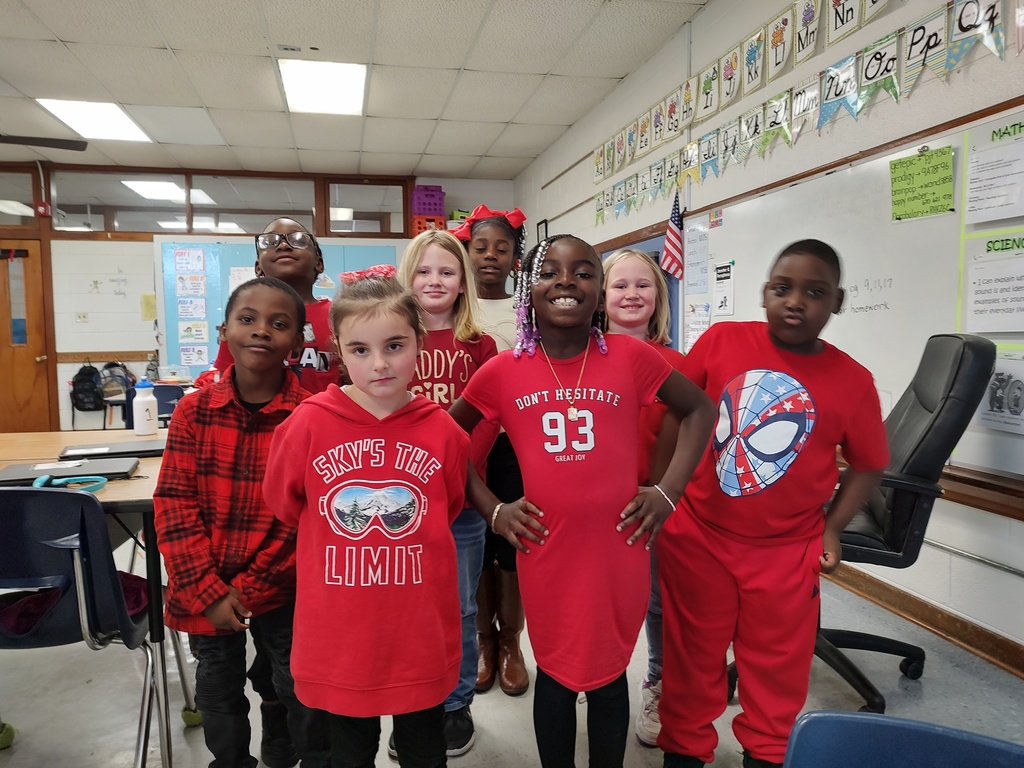 Two classroom groups of elementary students wearing red clothing for Red Ribbon Week. In the first image, a group of young children stands on a colorful classroom rug smiling in front of a smartboard