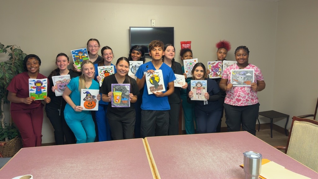 A group of Health Science students from West Bladen and East Bladen High Schools stand together in a classroom, each holding up a colorful fall-themed card they created for nursing home residents. The students are smiling and wearing scrubs in various colors, including blue, black, burgundy, and pink. The handmade cards feature pumpkins, scarecrows, and festive fall images, symbolizing their outreach project to share joy and kindness with local seniors.
