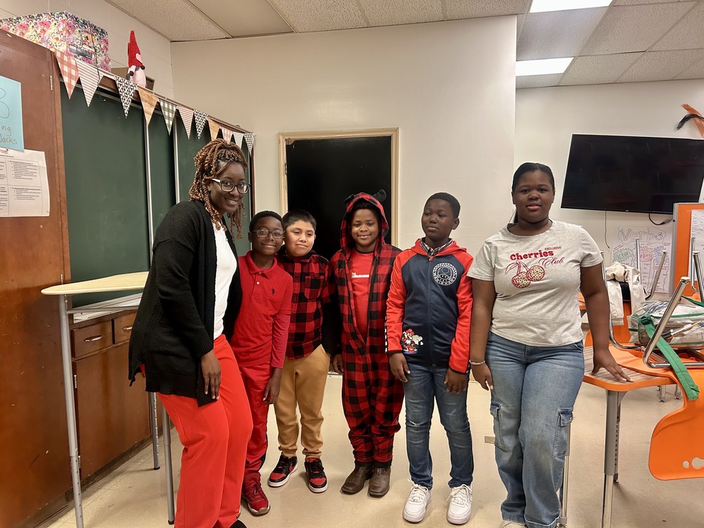 A teacher and five students stand together in a classroom decorated with banners and posters. Everyone is dressed in red for a themed spirit day, smiling as they pose by the chalkboard and desks.
