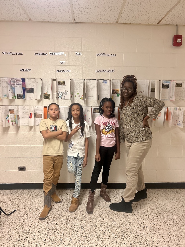 Three students and a teacher stand in a hallway at Elizabethtown Middle School. Behind them are student projects displayed on the wall titled “Ancient American Civilizations.” The group smiles proudly, showing off their work and school spirit.