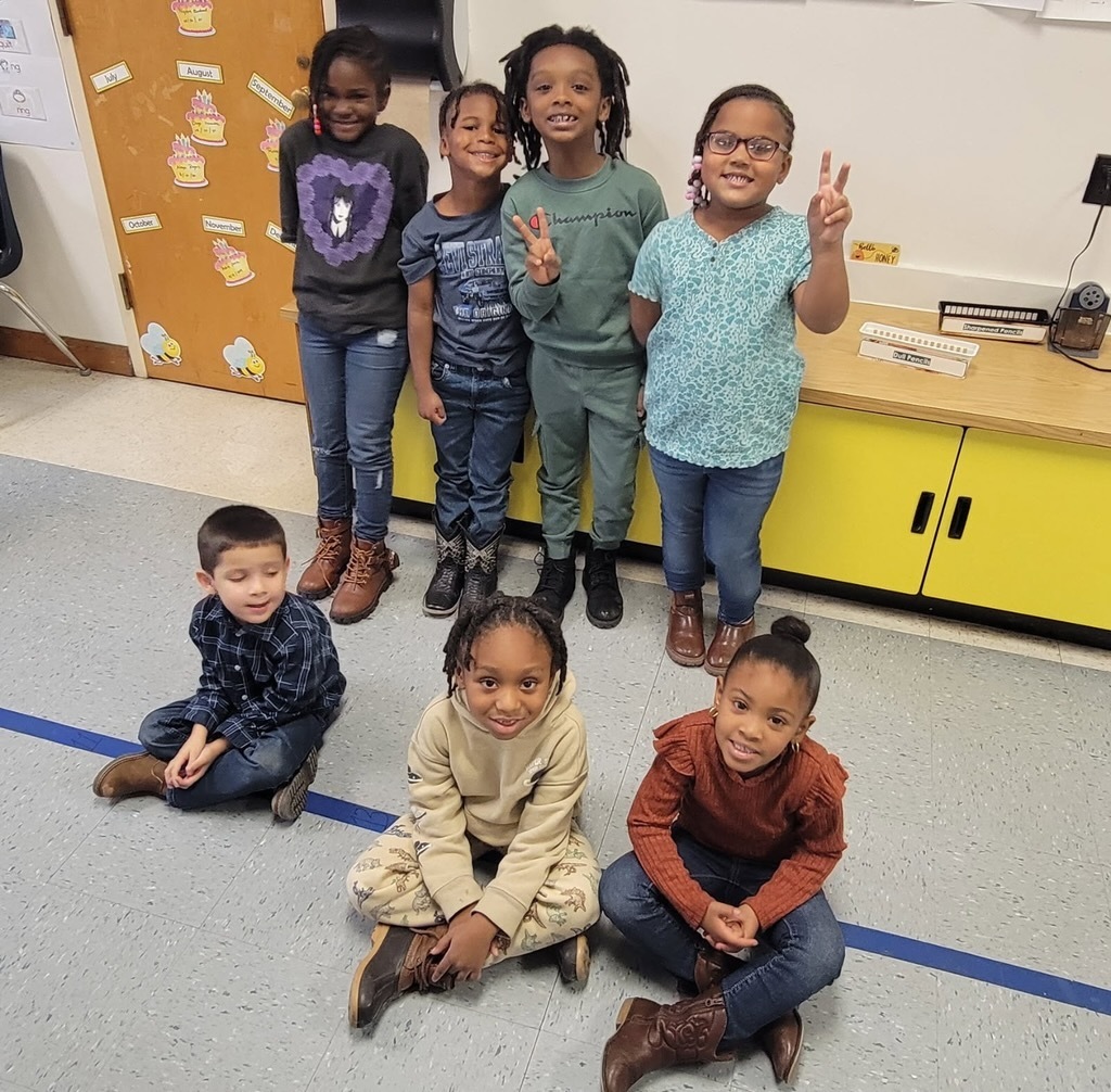 Seven students pose in a classroom — four standing and three sitting on the floor. Many are wearing cowboy boots or brown shoes, showing their Red Ribbon Week spirit.