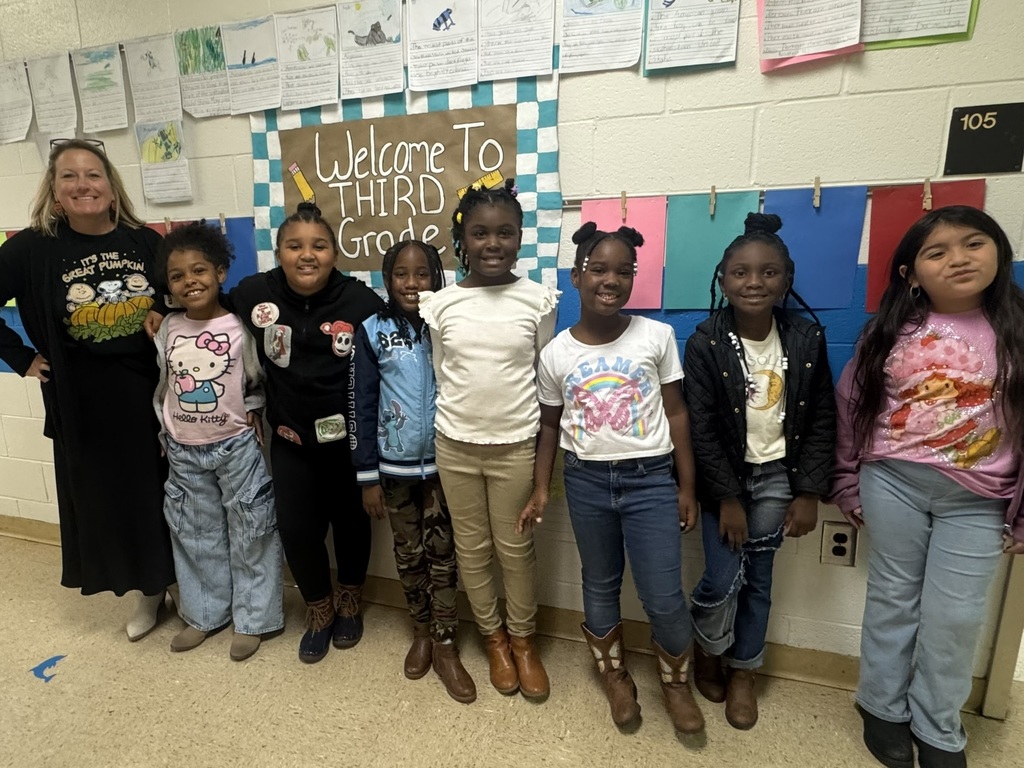 A teacher and seven students smile in front of a classroom wall that says “Welcome to Third Grade.” Students wear boots and fun shirts to match the Red Ribbon Week theme.