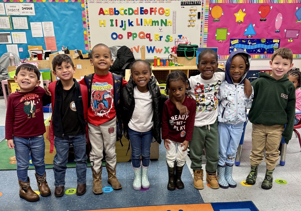Eight kindergarten students stand side-by-side in front of a colorful alphabet wall. They smile proudly, showing off their boots and sneakers.