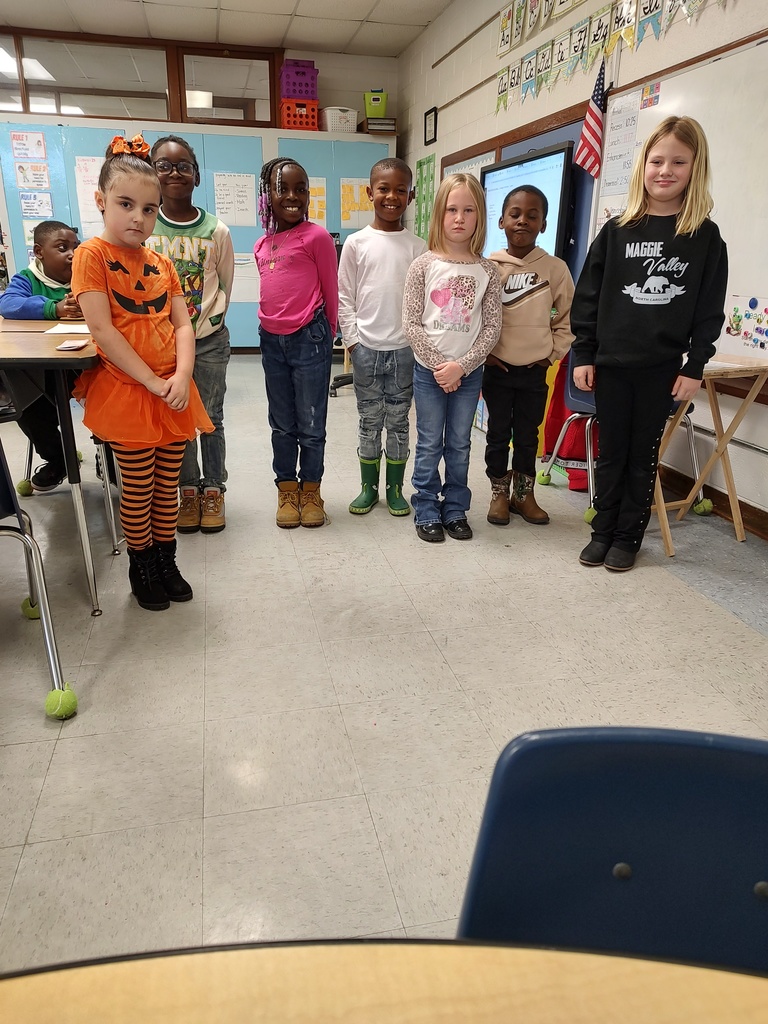 A group of seven smiling students stand together in a classroom, several wearing boots to celebrate Red Ribbon Week. One student is dressed in an orange pumpkin outfit with striped leggings.