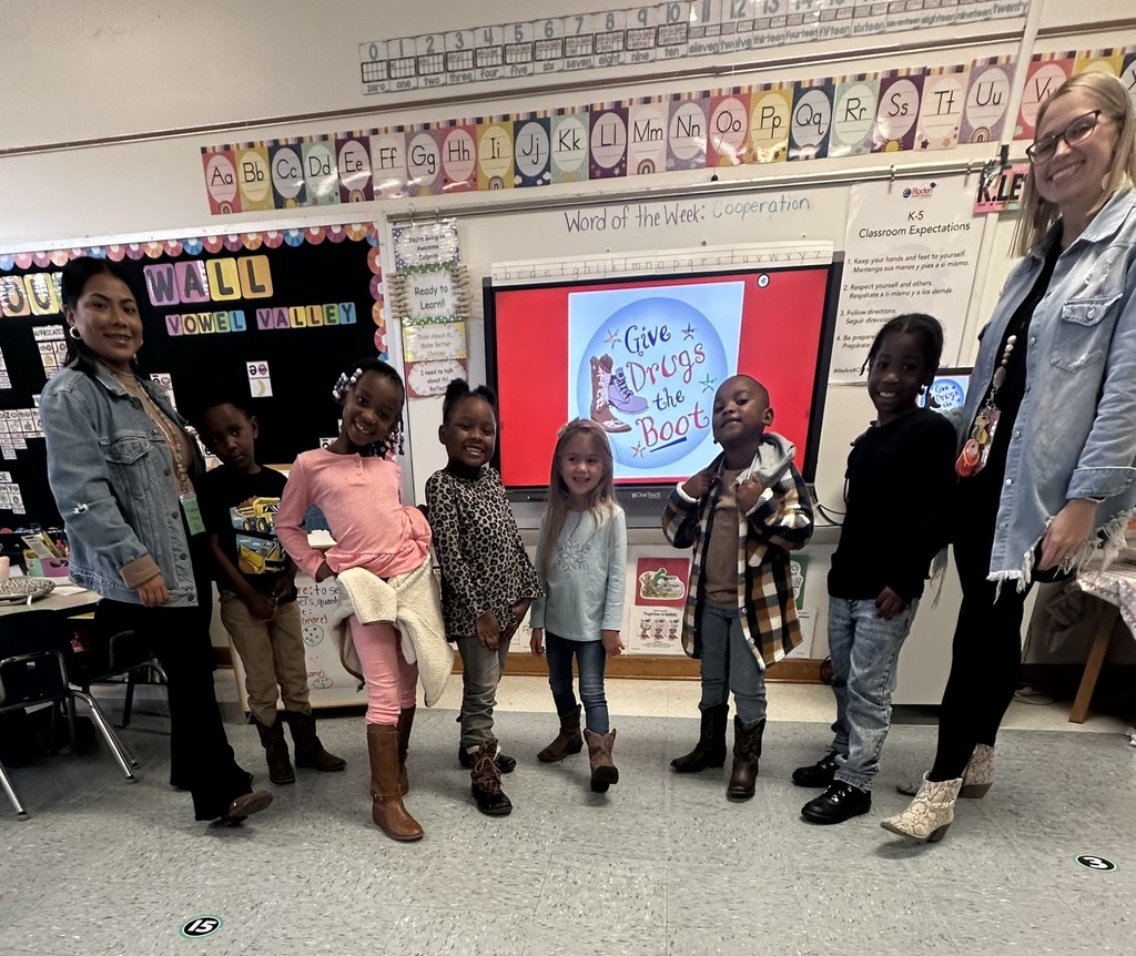 A group of young students and two teachers stand smiling in a classroom in front of a display screen that reads “Give Drugs the Boot.” The students are dressed in fall outfits and wearing boots as part of Red Ribbon Week. The classroom background includes colorful alphabet borders and a “Word of the Week: Cooperation” sign.