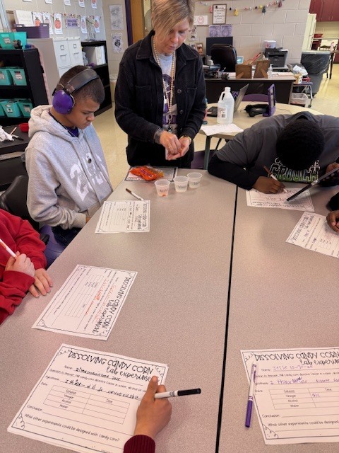A teacher works with a small group of high school students at a classroom table. The students are writing on lab sheets labeled “Dissolving Candy Corn” while discussing their results. A green drink bottle and a laptop are on the table, with posters and decorations visible in the background.
