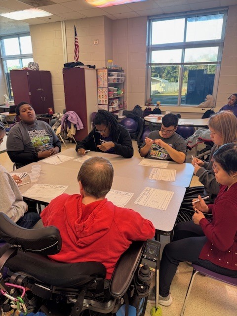 A teacher works with a small group of high school students at a classroom table. The students are writing on lab sheets labeled “Dissolving Candy Corn” while discussing their results. A green drink bottle and a laptop are on the table, with posters and decorations visible in the background.