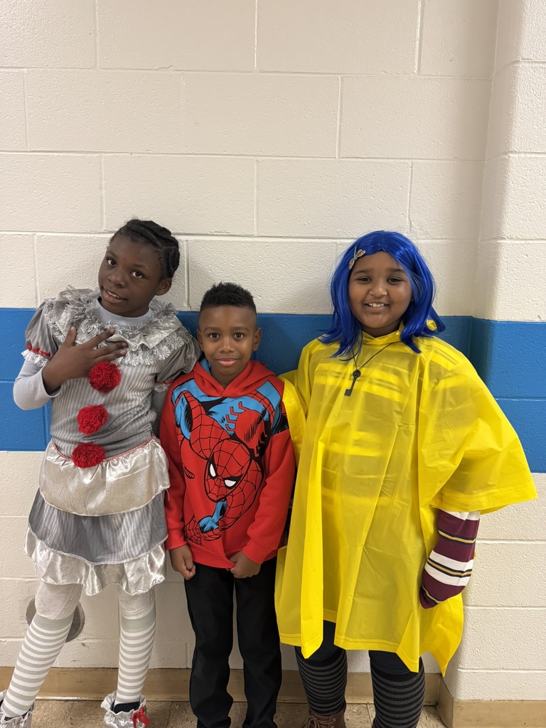 Three students in costumes—one in a gray clown outfit with red pom-poms, another in a red superhero hoodie, and one in a yellow raincoat and blue wig.