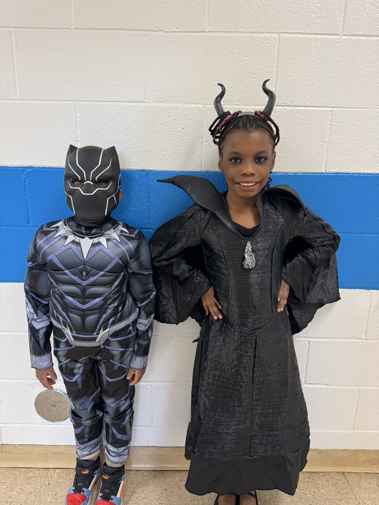 Two students dressed as a superhero and a dark fairy, smiling and standing against a blue and white wall.