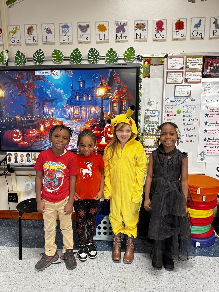 Four students smiling in a classroom decorated for fall; wearing themed outfits including a superhero shirt, Halloween leggings, a yellow onesie with ears, and a black layered dress.