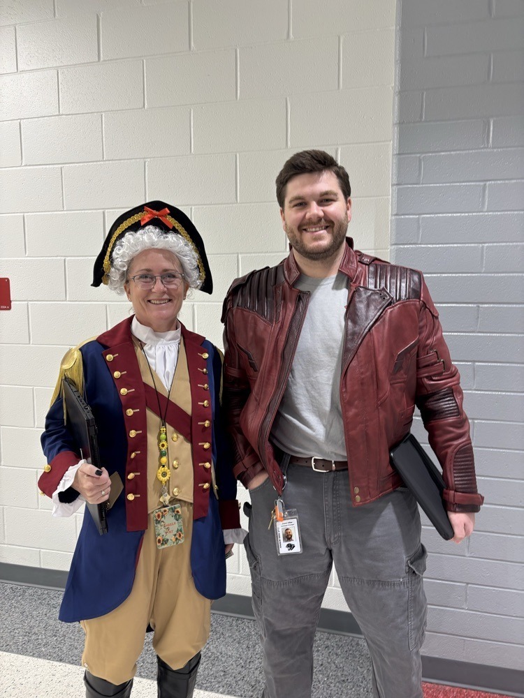 Two staff members smile in costumes—one in colonial attire and the other in a red jacket.