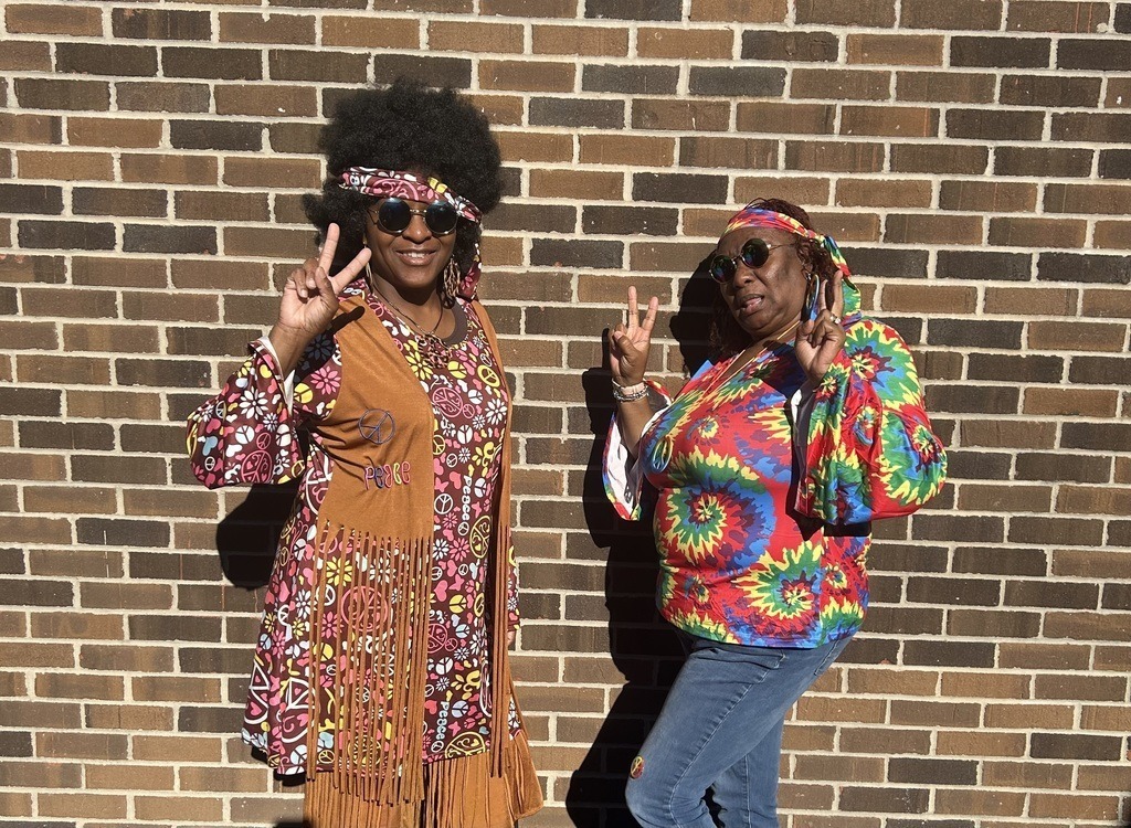 Two staff members dressed in colorful 70s outfits pose flashing peace signs.