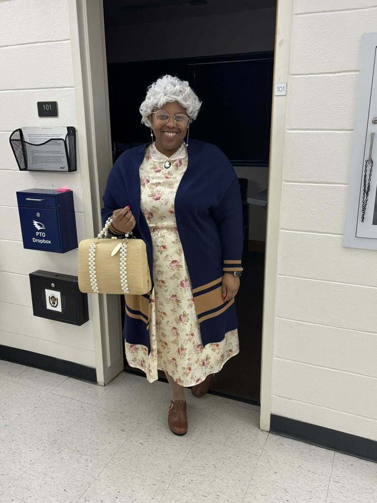 A staff member dressed as an elderly lady with gray wig, glasses, and floral dress smiles.