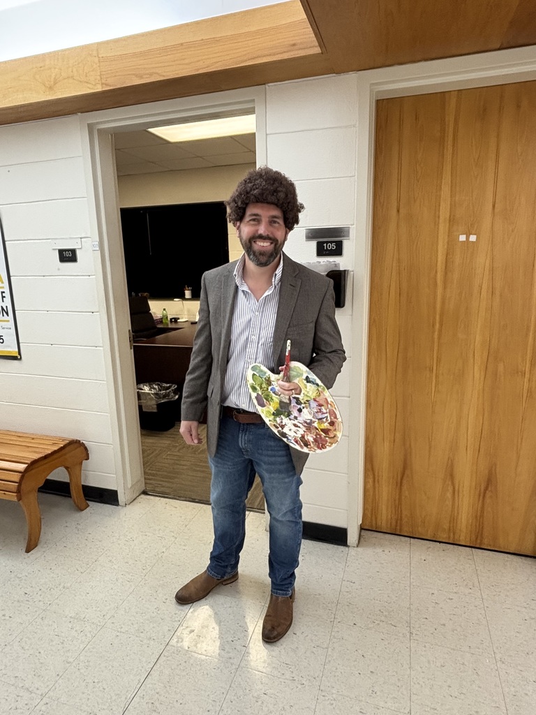 A staff member dressed as Bob Ross holds a paint palette and brush, smiling in the hallway.