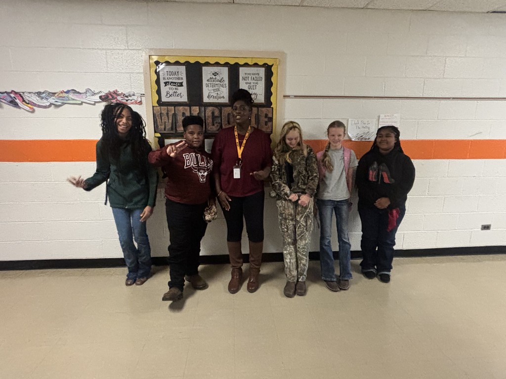 A group of students and a teacher stand together in a school hallway, smiling for a Red Ribbon Week photo. The bulletin board behind them features motivational messages and orange accents.