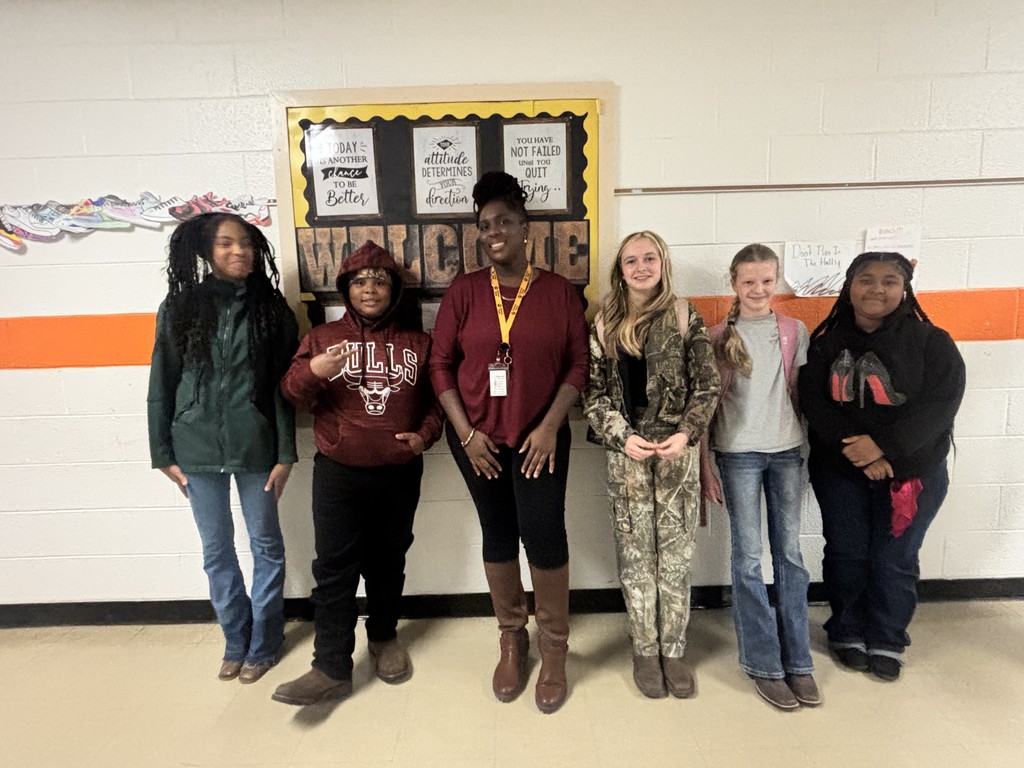 The same group of students and teacher stand lined up in a school hallway, smiling for a Red Ribbon Week photo in front of a bulletin board that says “Welcome.”