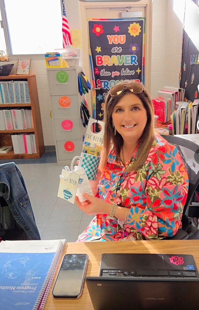 A smiling teacher sits at her desk holding a white mug filled with small treats and a tag that reads “I’ve Been Mugged.” She’s wearing a colorful floral blouse, and her classroom is decorated with bright, motivational posters.