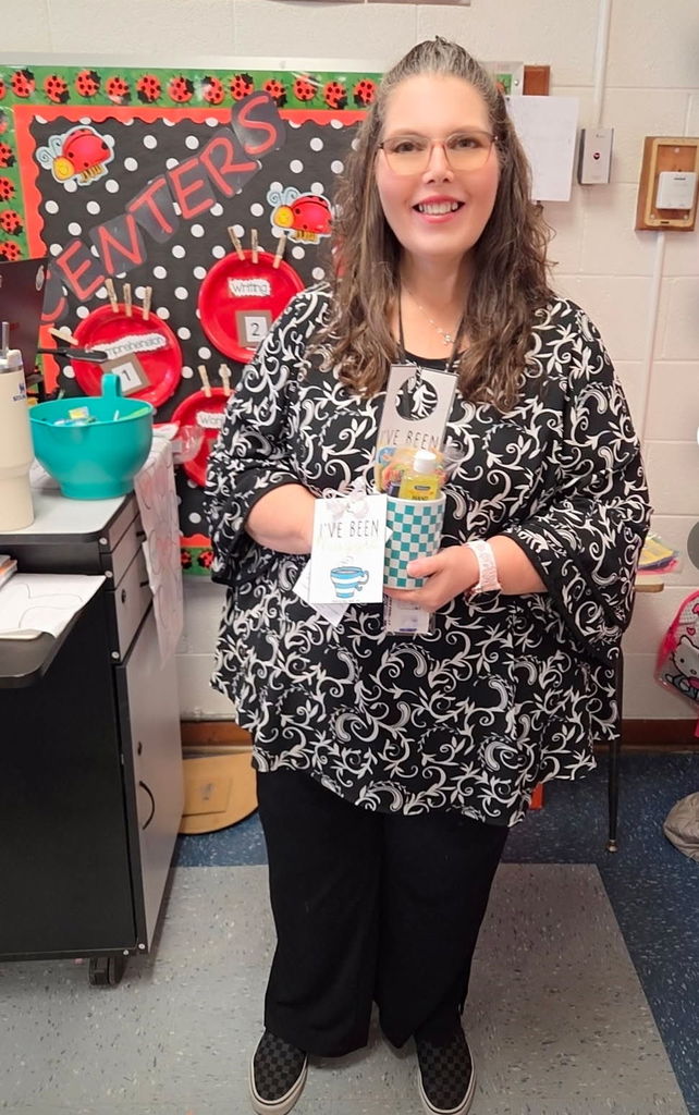 A teacher stands in front of a “Centers” bulletin board decorated with red ladybugs, holding a checkered blue “I’ve Been Mugged” mug filled with treats. She’s smiling and dressed in a black and white patterned top.