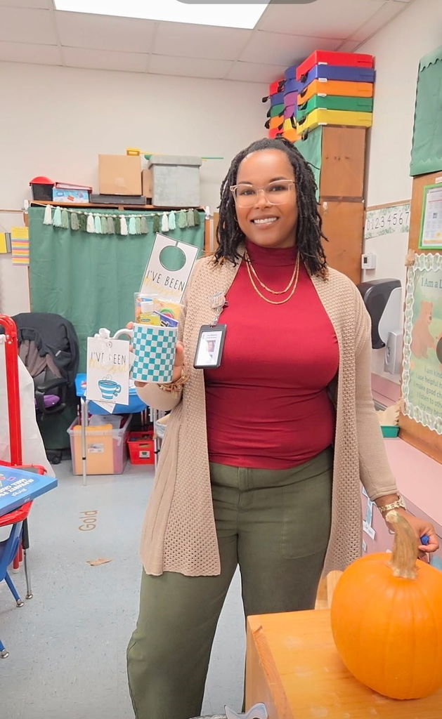 A smiling teacher stands in her classroom holding a “I’ve Been Mugged” mug filled with goodies. She’s wearing a red top with an open cardigan, and a pumpkin sits on the table beside her.