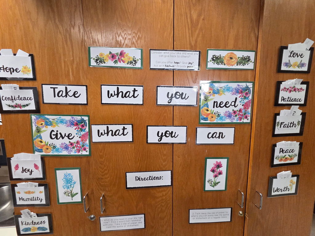A wooden cabinet display in the teacher lounge decorated with colorful floral signs and motivational words. The main message reads, “Take what you need, give what you can.” Around the display are small pockets labeled with uplifting words such as “Hope,” “Confidence,” “Joy,” “Humility,” “Kindness,” “Love,” “Patience,” “Faith,” “Peace,” and “Worth,” each containing slips of paper with inspirational quotes. The directions encourage staff to take a quote they need and share one to inspire others.